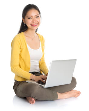 Young woman sits cross-legged on the floor with a laptop open in her lap, her fingers on the keyboard. She is looking at the camera and smiling.