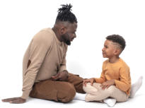 A dad and his son are sitting on the ground facing each other.  They are smiling and seem to be having a good conversation.
