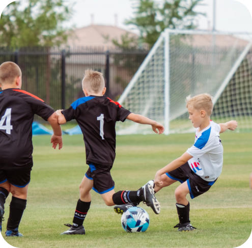 Three school-age boys in uniforms are playing soccer on a field with a goal in the background. Two of them, from different teams, are trying to kick the ball at the same time.