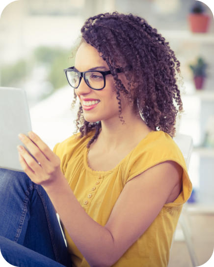 A young woman with glasses and casual clothing is seated and looking at a tablet that is propped on her knees. The woman is looking at the screen and smiling.