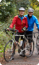 A middle aged couple in full biking gear are standing next to their bicycles on a path in the woods.