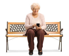 An elderly lady is sitting on a bench using her cell phone and smiling.