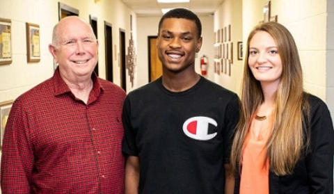 High school football player Taevone Johnson stands in the hallway of his school, between assistant coach Chuck Mardis and athletic trainer Lindsey Braddock