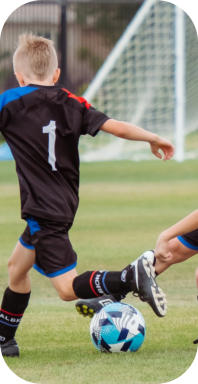 Three school-age boys in uniforms are playing soccer on a field with a goal in the background. Two of them, from different teams, are trying to kick the ball at the same time.