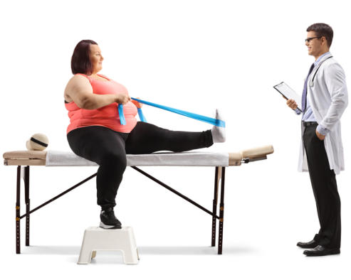 A middle-aged woman in athletic wear sits on a physical therapy table. She has one leg extended on the table and is holding a resistance band with both hands as it pulls back the top of her foot in a stretch. Her doctor looks on, standing with a clipboard