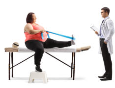 A middle-aged woman in athletic wear sits on a physical therapy table. She has one leg extended on the table and is holding a resistance band with both hands as it pulls back the top of her foot in a stretch. Her doctor looks on, standing with a clipboard