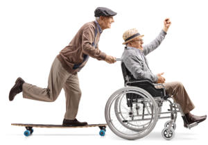 An old man riding a skateboard is pushing another old man in a wheelchair. They are both wearing jaunty hats and seem to be having a lot of fun.