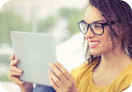 A young woman with glasses and casual clothing is seated and looking at a tablet that is propped on her knees. The woman is looking at the screen and smiling.