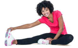 A young woman in athletic wear is sitting on the floor stretching her right leg and touching her toes.