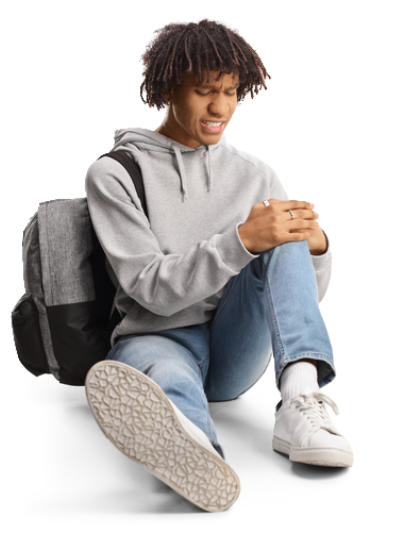 A young man in jeans and a sweatshirt is sitting on the ground wearing a backpack and holding his knee in pain.