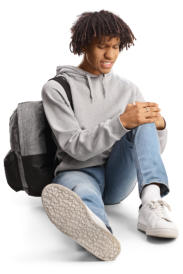 A young man in jeans and a sweatshirt is sitting on the ground wearing a backpack and holding his knee in pain.