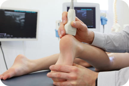 A medical technician uses an ultrasound wand to examine the heel of a young patient. The ultrasound screen image is in the background.