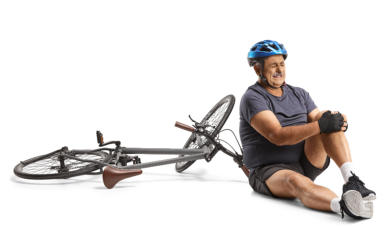 A senior man in athletic clothing and a bicycle helmet sits on the ground holding his knee and grimacing. His bike is lying on the ground next to him.