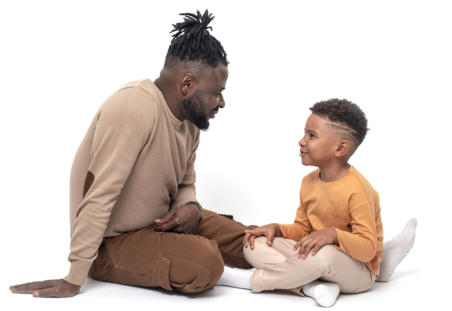 A dad and his son are sitting on the ground facing each other.  They are smiling and seem to be having a good conversation.