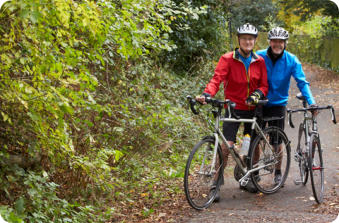 A middle aged couple in full biking gear are standing next to their bicycles on a path in the woods.